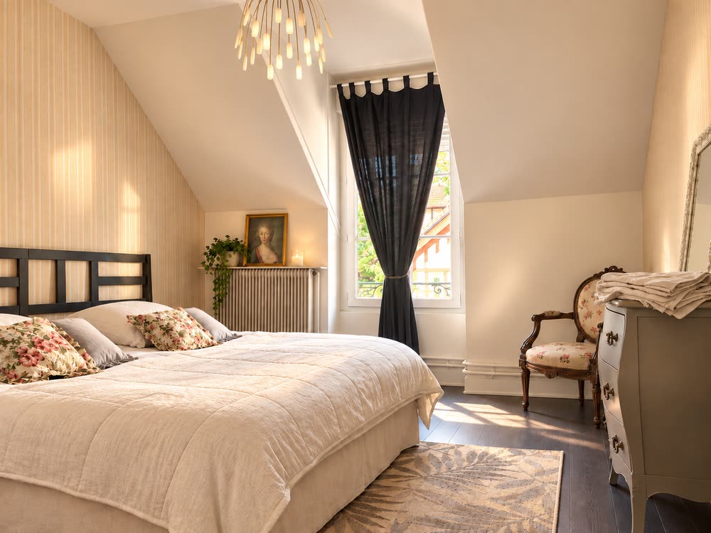 Une chambre du gîte sous la pente du toit, lit double à tête de lit sombre et fauteuil près de la fenêtre
